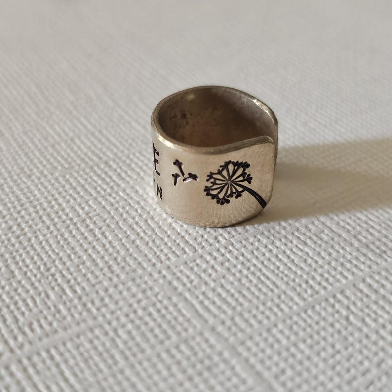 May include: A silver-colored ring with a dandelion design and the word "Believe" etched on the band. The ring has a wide band and a slightly textured surface, suitable for everyday wear.