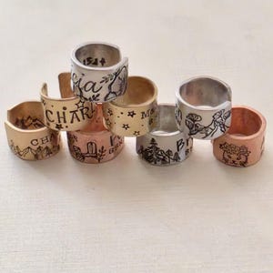 May include: A collection of handmade metal rings in various colors, including gold, silver, and copper. Each ring features unique engraved designs, such as mountains, floral patterns, and text. The rings are stacked, showcasing their individual details.