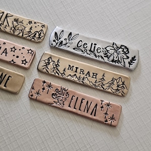 May include: Close-up of several metal name tags with stamped designs and names. One tag features a deer and mountain scene, another has the name "Celie" with floral accents, and a third reads "Mirah" with a mountain and tree design.