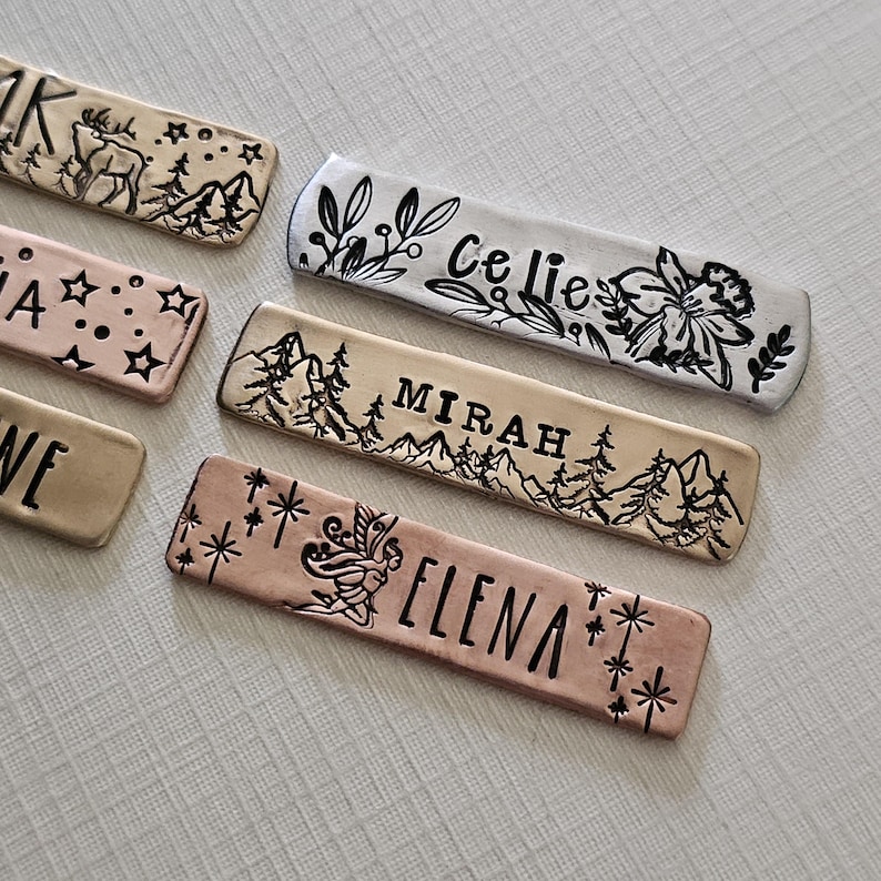 May include: Close-up of several metal name tags with stamped designs and names. One tag features a deer and mountain scene, another has the name "Celie" with floral accents, and a third reads "Mirah" with a mountain and tree design.