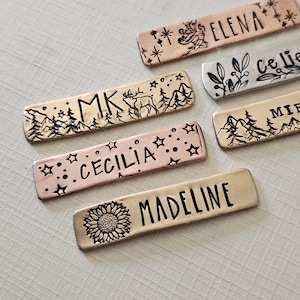 May include: Close-up of several rectangular metal name tags. Each tag features a name, such as "Elena" or "Cecilia", and decorative elements like stars, a sunflower, or mountain and forest designs. The tags have a rustic, handmade appearance.