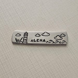 May include: A silver-colored metal bar with a lighthouse, clouds, and waves design. The word "ALENA" is stamped in the center. The bar is rectangular with rounded edges, and the design is etched into the metal.