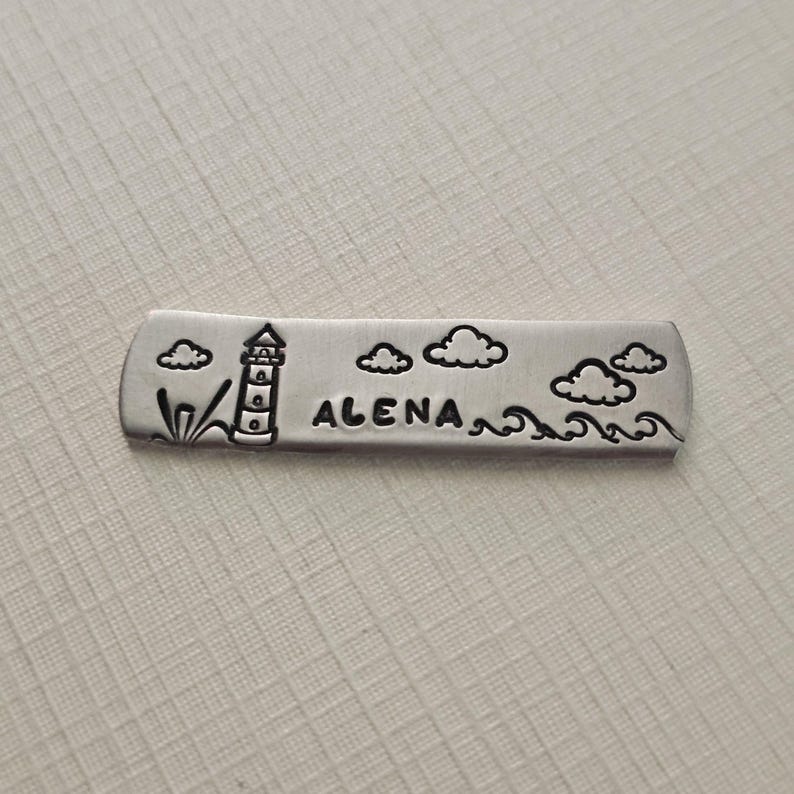May include: A silver-colored metal bar with a lighthouse, clouds, and waves design. The word "ALENA" is stamped in the center. The bar is rectangular with rounded edges, and the design is etched into the metal.