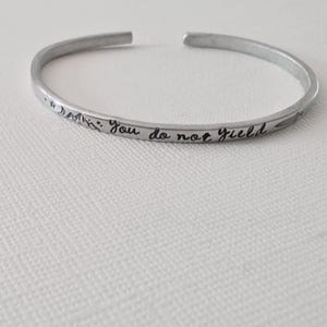 May include: A silver-toned metal cuff bracelet featuring an open design. The exterior surface is engraved with the text "You do not yield" in a cursive script, accompanied by small etched icons of a star, a crescent moon, mountains, and a sword.