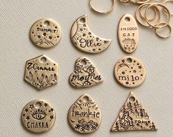 Puede incluir: Una colección de etiquetas de metal en tono dorado con diversas formas, incluyendo círculos, una luna creciente, un hexágono, un corazón y un triángulo. Cada etiqueta presenta diseños grabados y nombres como Summit, Ollie, Indoor Cat, Winner, Meimer, Milo, Chakra, Frankie y Frank.