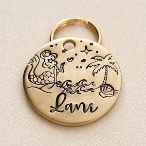 May include: A gold-colored, round pet tag with a mermaid, palm tree, and waves design. The name "Lani" is written in a cursive font. The tag has a gold ring for attachment.