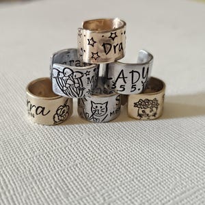 May include: A stack of handmade metal rings with various designs and text. One ring has stars and the word "Dra", another has a cat illustration, and others feature floral and abstract designs. The rings are in gold and silver tones.