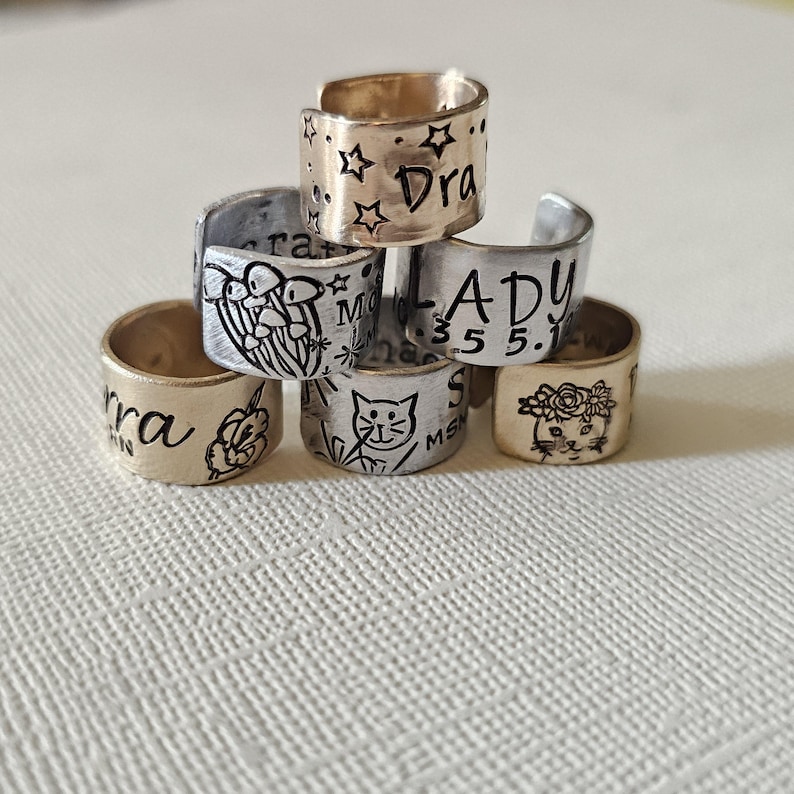 May include: A stack of handmade metal rings with various designs and text. One ring has stars and the word "Dra", another has a cat illustration, and others feature floral and abstract designs. The rings are in gold and silver tones.