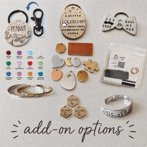 May include: A collection of metal pet tags, including round, oval, and bow-tie shapes with engraved text like PEANUT and CHIPPED, various metal charms, two silver-tone cuff bracelets, a birthstone color chart, and a small plastic polish kit.