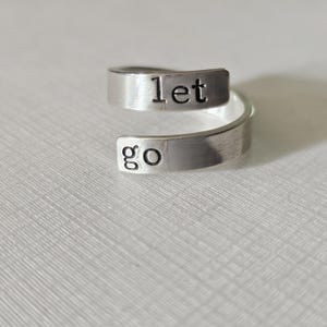 May include: A silver ring with the words "let go" engraved on it. The ring is designed to be worn on the finger with the words facing outward.