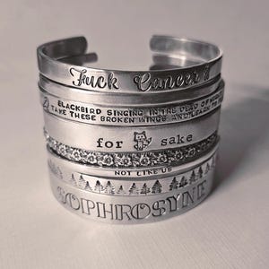 May include: A collection of silver metal cuff bracelets, each engraved with text. The bracelets feature phrases like "Fuck Cancer" and song lyrics. Some have floral or forest designs. The bracelets are stacked together.