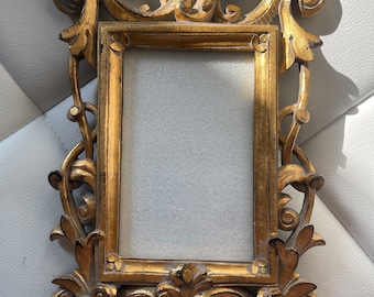 May include: A gold-colored ornate picture frame with a detailed, intricate design. The frame has a rectangular opening for a picture.