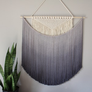 May include: A macrame wall hanging with a white triangle design and a gray ombre fringe. The fringe is made of yarn and is tied to a wooden dowel.
