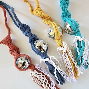 May include: Four macrame disco ball ornaments in shades of rust, blue, yellow, and teal. Each ornament features a mirrored disco ball, knotted cord, and a tassel of coordinating yarn and lace.