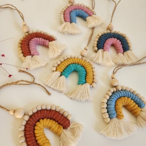 Könnte beinhalten: Fünf bunte, regenbogenförmige Wandbehänge aus Garn und Holzperlen. Jeder Regenbogen hat eine andere Farbkombination, darunter Rosa, Blau, Türkis, Gelb und Braun.