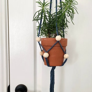 May include: A blue macrame plant hanger with wooden beads holds a terracotta pot with a green plant. The hanger is hanging from a white door.