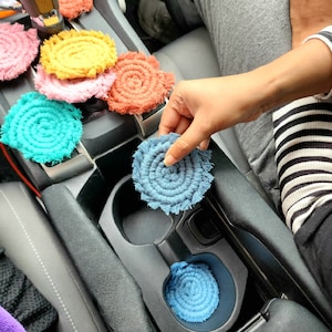 Puede incluir: Un juego de posavasos de crochet de colores con flecos. Los posavasos están en un coche, colocados en los portavasos.
