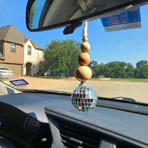 May include: A car interior with a rearview mirror and a hanging car charm. The charm is a disco ball with a string of wooden beads in beige and brown.