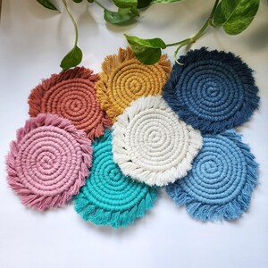 May include: Set of seven colorful crocheted coasters with fringe. The coasters are in shades of pink, blue, teal, white, and yellow.