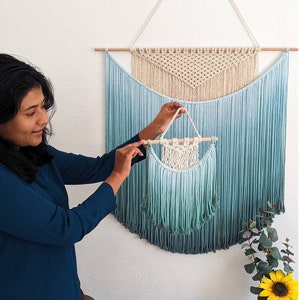 May include: A teal ombre macrame wall hanging with a smaller teal ombre macrame hanging in front of it. The larger hanging has a white macrame triangle in the center. The text "Color: Ombre Teal" is at the bottom of the image.