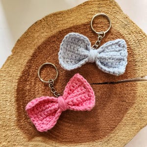 May include: Two handmade crochet bow keychains. One is light blue, the other is pink. Each bow is attached to a silver-colored keyring. The keychains are displayed on a textured, light brown wooden surface.