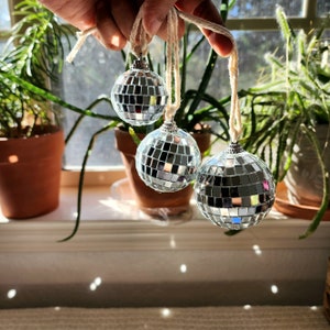May include: Three silver disco ball ornaments hanging from white string. The ornaments are round and have a mirrored surface.