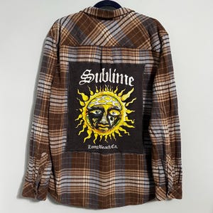 May include: Brown, blue, and grey plaid flannel shirt with a black patch featuring the word "Sublime" above a yellow sun graphic with a face. The text "Long Beach Ca." is below the sun.