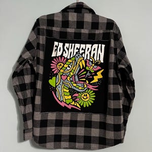 May include: A gray and black plaid flannel shirt with a large black patch on the back featuring the text "ED SHEERAN" above a colorful graphic of a snake, flowers, and lightning bolts.