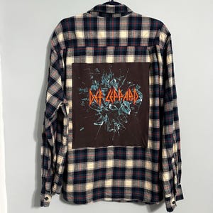 May include: Plaid flannel shirt in navy, cream, and red. The back features a brown patch with the Def Leppard logo and a shattered glass design. The shirt has long sleeves and a button-down front.