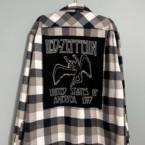 May include: Black, white, and gray plaid flannel shirt with a large black patch on the back. The patch features the text "LED-ZEPPELIN" above a graphic of a winged figure and the text "UNITED STATES OF AMERICA 1977".
