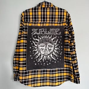 May include: Yellow, black, and white plaid flannel shirt with a large black square on the back. The square features the word "SUBLIME" above a white sun graphic with a face. The shirt has a button-down front and long sleeves.