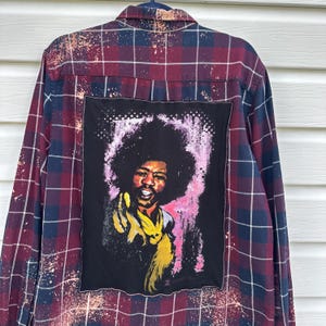 May include: A long-sleeved, button-down flannel shirt in a burgundy, navy, and white plaid pattern. The back features a black patch with a colorful portrait of a musician. The shirt has a distressed, vintage look with paint splatters.