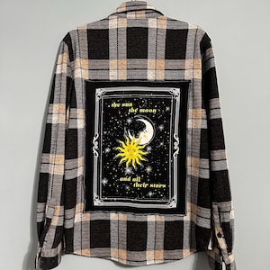 May include: A long-sleeved flannel shirt with a plaid pattern in shades of brown, tan, and black. The back features a black square with a celestial design of the sun, moon, and stars, with the text ".the sun the moon and all their stars."