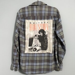 May include: A button-down flannel shirt in a plaid pattern of gray, blue, and green. The back features a rectangular patch with a black and white image of Whitney Houston and the text "WHITNEY HOUSTON" in orange.