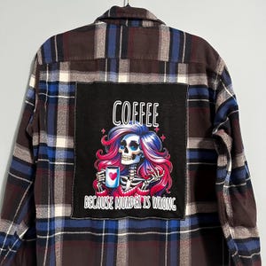 May include: A plaid flannel shirt in shades of brown, blue, and white. The back features a black patch with the word "COFFEE" above a skeleton graphic with pink and purple hair, holding a mug. The text "BECAUSE MURDER IS WRONG" is below.