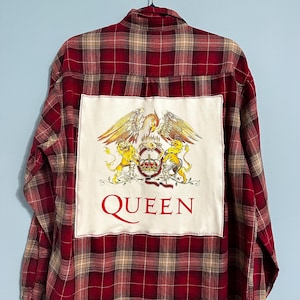 May include: Red and brown plaid button-down shirt with a large white patch on the back. The patch features a gold and red emblem with the word "QUEEN" in red block letters. The shirt is hanging on a hanger.