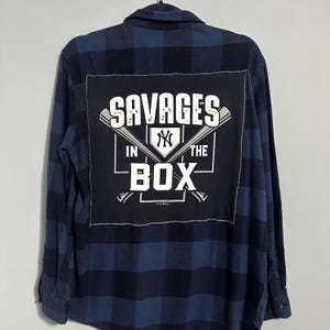 May include: A blue and black plaid flannel shirt with a large black patch on the back. The patch features the text "SAVAGES IN THE BOX" with baseball bats and the New York Yankees logo in white.