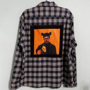 May include: A long-sleeved, button-down shirt with a black and navy blue plaid pattern. The back of the shirt features a square patch with an image of a person wearing glasses and a hat, holding a flower, set against an orange background.