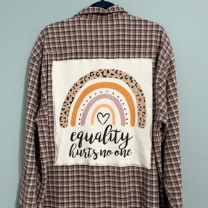 May include: A brown and white plaid long-sleeve shirt featuring a large white rectangular patch on the back. The patch displays a colorful rainbow with a leopard print arch, a small black heart, and the text equality hurts no one in black.