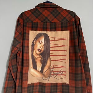 May include: A long-sleeved, button-down shirt with a red and brown plaid pattern. The back of the shirt features a rectangular patch with a portrait of Aaliyah and her signature, with the name "AALIYAH" printed vertically.