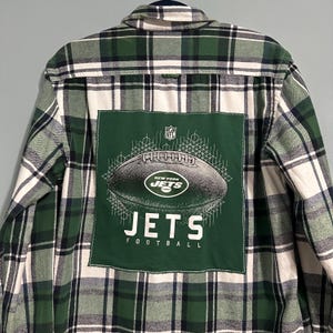 May include: Green, white, and navy blue plaid flannel shirt. The back features a green patch with a football graphic and the words "NEW YORK JETS" and "JETS FOOTBALL".