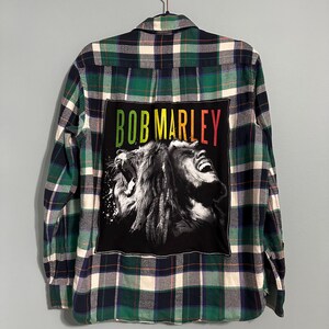 May include: Green, blue, and white plaid flannel shirt with a black patch featuring Bob Marley's name in green, yellow, and red. The patch displays a black and white image of a lion's face.