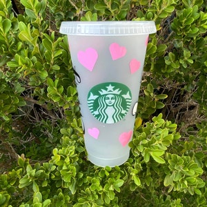 Puede incluir: Un vaso fr&iacute;o de Starbucks de pl&aacute;stico transparente con tapa, decorado con pegatinas de corazones rosas. El logotipo de Starbucks es visible en la parte delantera. La taza est&aacute; colocada sobre un fondo de follaje verde.