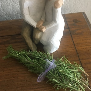 May include: A bundle of fresh rosemary tied with a lavender ribbon, resting on a wooden surface. A white ceramic sculpture of a couple embracing is in the background.