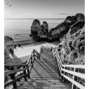 Pode incluir: Uma escada de madeira com corrim&atilde;os leva a uma praia de areia com penhascos rochosos e o oceano ao fundo.