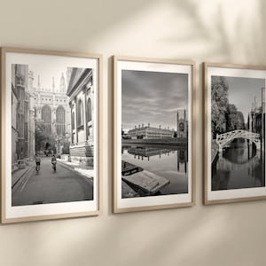 May include: Three black and white framed photographs of Cambridge, England. The first photo shows a narrow street with two people riding bicycles. The second photo shows a river with a rowboat and a building in the background. The third photo shows a bridge over a river with trees on the banks.
