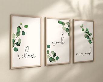May include: A set of three vertical wall art prints in light wood frames. Each white print features green eucalyptus branches and black cursive text. The first print says relax, the second says soak, and the third says unwind.