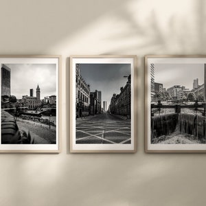 May include: Three black and white framed photographs of cityscapes. The first photo shows a view of a city with a tall building and a bridge. The second photo shows a street with buildings on either side. The third photo shows a canal with a waterfall.