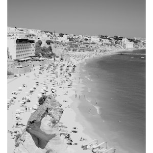 Pode incluir: Uma fotografia a preto e branco de uma praia com um penhasco rochoso em primeiro plano. A praia est&aacute; cheia de pessoas e guarda-s&oacute;is. H&aacute; edif&iacute;cios no fundo.