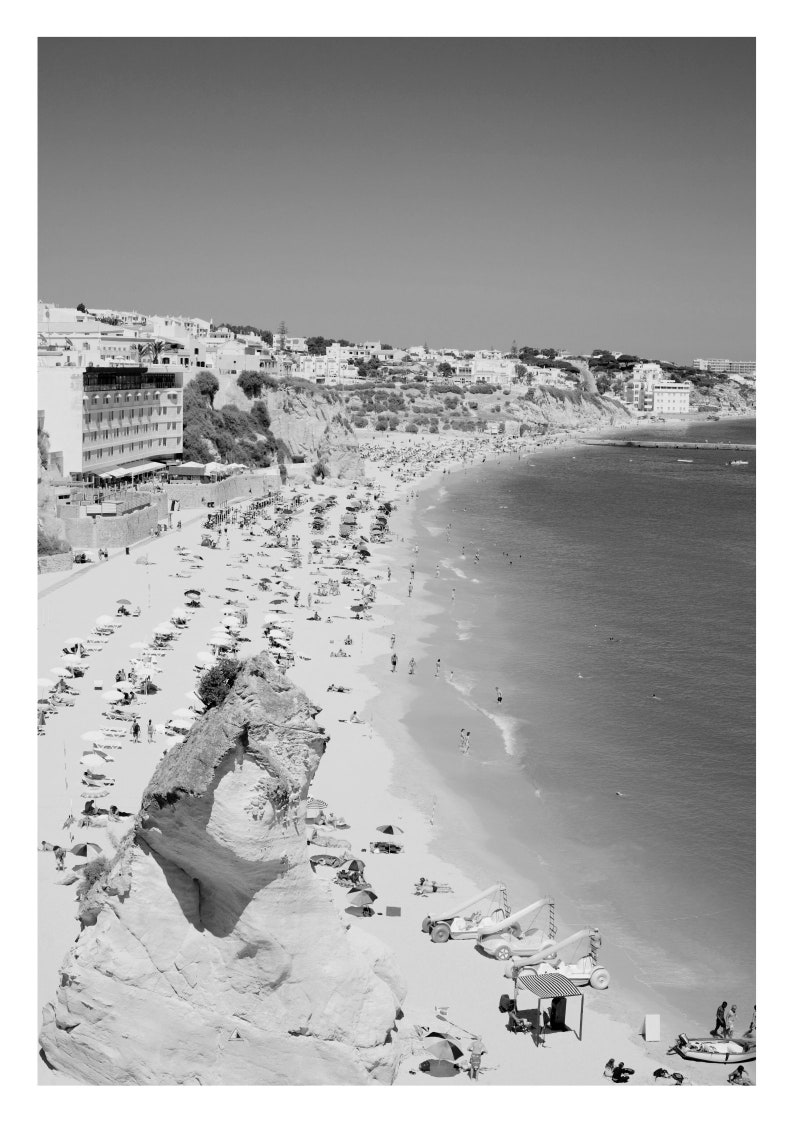 Pode incluir: Uma fotografia a preto e branco de uma praia com um penhasco rochoso em primeiro plano. A praia est&aacute; cheia de pessoas e guarda-s&oacute;is. H&aacute; edif&iacute;cios no fundo.